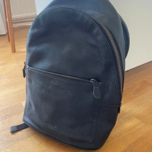 Svart ryggsäck från Coach - Snygg svart ryggsäck från Coach i skinn med ordentlig dragkedja. Perfekt för både vardag och resa. Rymlig design med justerbara axelremmar och en praktisk ytterficka med dragkedja. Köpt för några år sedan och aldrig använd. Nypris verkar vara mellan 4000-5000
