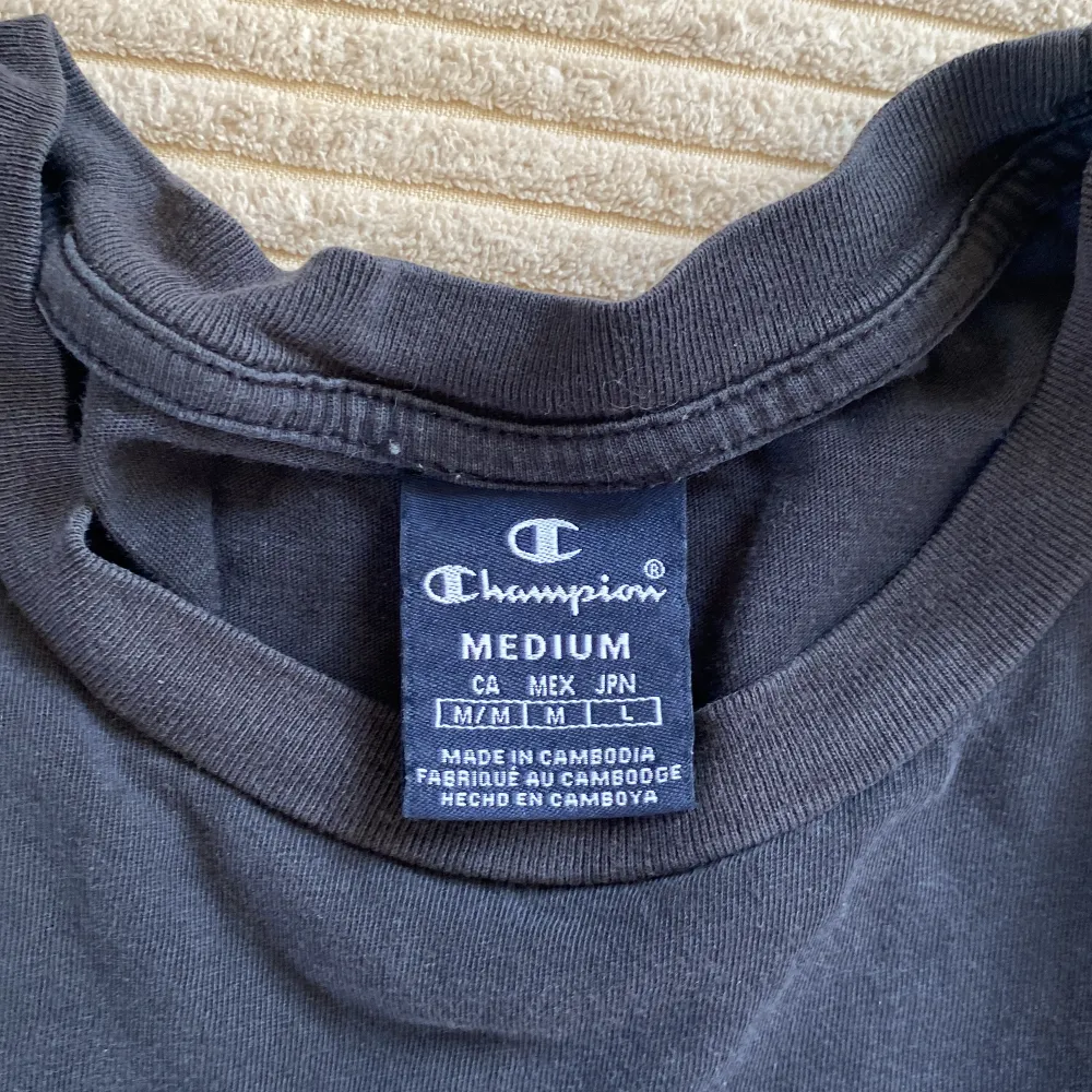 Svart t-shirt från Champion med vit logga på bröstet och loggoband längs axlarna. Klassisk passform och korta ärmar. Perfekt för en sportig och avslappnad stil.. T-paidat.
