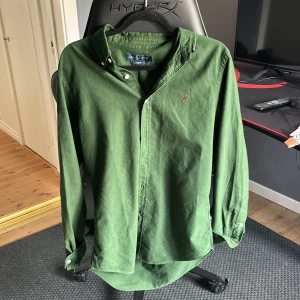 Grön skjorta från Ralph Lauren - Säljer en grön skjorta från Ralph Lauren i slim fit-modell. Skjortan har klassisk krage med knappar, lång ärm och den ikoniska broderade loggan på bröstet. Perfekt för dig som gillar stilrena plagg med snygga detaljer. Loggan på bröstet är målar från orange till svart annars 10/10
