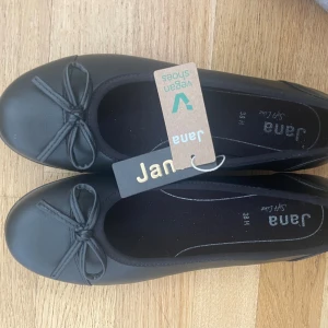 Svarta ballerinaskor med rosett från Jana - Säljer ett par svarta ballerinaskor från Jana med söt rosettdetalj framtill. Skorna är veganska och har en klassisk rund tå. 