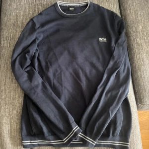Mörkblå tröja från Hugo Boss - Snygg mörkblå tröja från Hugo Boss med diskret logga på bröstet. Tröjan har rund halsringning och ribbade muddar med vita ränder vid ärmslut och nederkant. Perfekt för en stilren och avslappnad look. St L men är som M.