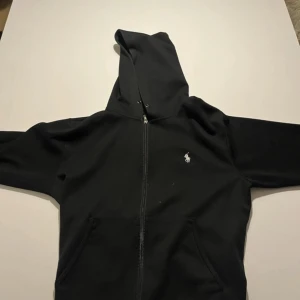 Ralph Lauren zip hoodie - Svart hoodie från Polo Ralph Lauren med dragkedja och klassisk vit broderad logga på bröstet. Är använd men fortfarande i bra skick. Skriv om ni undrar något