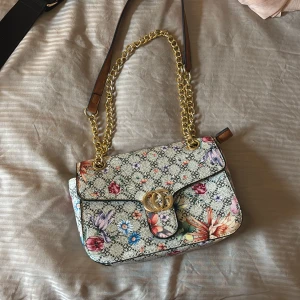 Blommig väska - Superfin axelväska  med blommigt mönster och guldfärgad kedja. Väskan har GG-logga framtill, läderdetaljer och innerfack med dragkedja. Perfekt för dig som vill ha en färgglad och trendig accessoar.