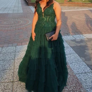 Mörkgrön långklänning med spets och volanger - Säljer en elegant mörkgrön långklänning med djup v-ringning och spetsdetaljer upptill. Kjolen har flera lager volanger i tyll som ger en lyxig känsla. Perfekt för bal eller fest! Klänningen har smala axelband och markerad midja. Storlek 38/40