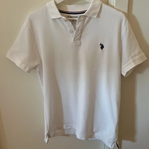 Vit pikétröja från U.S. Polo Assn. - Säljer en klassisk vit pikétröja från U.S. Polo Assn. med broderad logga på bröstet. Tröjan har korta ärmar, krage och knappar framtill. Perfekt för en stilren och sportig look. Inga defekter. Tveka inte att höra av dig vid funderingar.