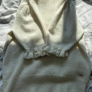 Vit stickad tröja från Holly & Whyte - Säljer en söt vit stickad tröja från Holly & Whyte. Tröjan har volangdetaljer vid hals och ärmslut, samt dekorativa knappar på ärmarna. Mjuk och mysig med fint mönsterstickat tyg och hög hals.