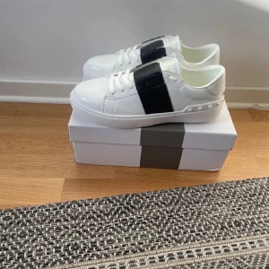 Valentino svart opens - Säljer ett par snygga vita sneakers från Valentino med en bred svart rem över sidan och små nitar längs sulan. Skorna har klassisk rund tå och är perfekta för dig som gillar stilrena detaljer.