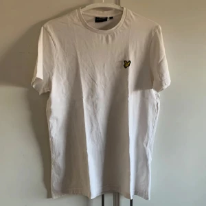 Vit t-shirt från Lyle & Scott - Säljer en stilren vit t-shirt från Lyle & Scott med en liten broderad logga på bröstet. T-shirten har en klassisk rund hals och korta ärmar. Perfekt för en avslappnad look!