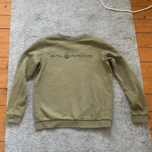 Olivgrön sweatshirt från Sail Racing - Säljer en olivgrön sweatshirt från Sail Racing med tryckt logga på bröstet. Tröjan har rund halsringning och långa ärmar. Tillverkad i en mjuk bomulls- och polyestermix för extra komfort.