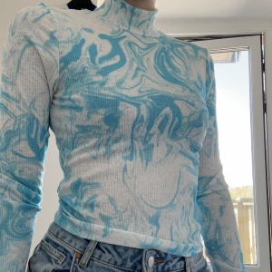 Blåvit ribbad polotopp med mönster - Säljer en ribbad polotopp i vitt med blått swirl-mönster. Tröjan har lång ärm och hög hals, sitter tight och är tillverkad i stretchigt material. Perfekt att styla med jeans för en cool look. Den är ganska tunn så den kan absolut bäras på sommaren. 