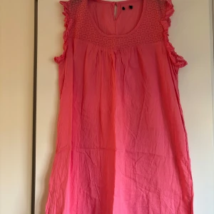 Rosa kortklänning/tunika med volang och spets - Säljer en söt rosa kortklänning/tunika med volangdetaljer på axlarna och spetsmönster upptill. Klänningen är ärmlös och har en rund halsringning med knapp i nacken. Perfekt för varma dagar och somriga tillfällen.