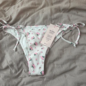 Vit blommig bikiniunderdel från Nelly - Köptes för 179! Supersöt bikiniunderdel från Nelly i vitt med små rosa blommor och gröna blad. Modellen har smala knytband i sidorna för perfekt passform och en klassisk, lite högre skärning. 