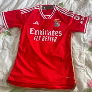Benfica hemma matchtröja Adidas S  - Snygg äkta Benfica fotbollströja från Adidas i rött med vita detaljer och klubbmärke på bröstet. Helt oanvänd med prislappen kvar. Tröjan köptes på plats i Benficas supportshop på Sport Lisboa e Benfica Stadium. Köpte tröjan för 100€ vilket motsvarar ca 1100kr 