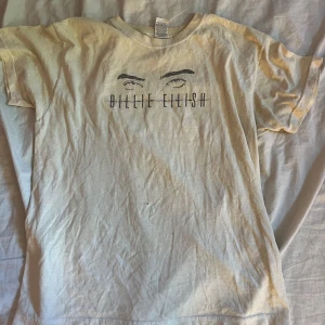 Billie Eilish t-shirt i beige bomull - Säljer en beige t-shirt med tryck av Billie Eilishs ögon och namn på bröstet. T-shirten är i mjuk bomull och har klassisk passform med korta ärmar och rund hals. Den har ett pyttelitet hål rakt på magen som man knappt ser. Men den är jättefin och passar bra.