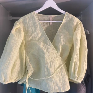 Ljusgul omlottblus med puffärm - Superfin ljusgul omlottblus från H&M med puffiga långa ärmar och knytband i sidan. Blusen har en lätt skrynklig struktur och v-ringning, perfekt för dig som gillar en luftig och trendig look. Materialet känns svalt och passar grymt till vår och sommar.