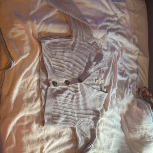 Ljusgrå ribbad kofta från H&M - Superfin ljusgrå kofta från H&M i ribbstickad bomull. Modellen är croppad med v-ringning och stora knappar framtill. Perfekt att slänga över en topp för en chill look. Långa ärmar.