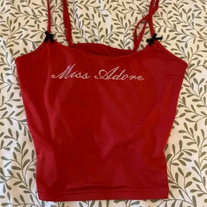 Röd linne Miss Adore med strass - Supersöt röd linne med smala axelband från Miss Adore. Texten på bröstet är dekorerad med glittrande strass och det finns små svarta rosetter vid axelbanden. Perfekt för dig som vill sticka ut lite extra.