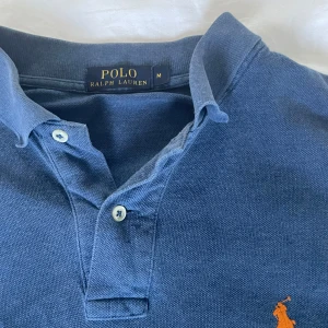 Blå pikétröja från Ralph Lauren - Klassisk blå pikétröja från Polo Ralph Lauren med korta ärmar och krage. Tröjan har två knappar vid halsen och den ikoniska orange broderade loggan på bröstet. Perfekt för en clean och avslappnad stil. Köpt för 1500 kr på Zalando• Skick 9/10• Priset är inte hugget i sten, så kom med bud!