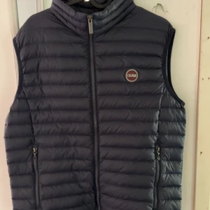 Mörkblå dunväst från Colmar - Snygg mörkblå dunväst från Colmar med quiltad design och hög krage. Västen har två dragkedjefickor framtill och stängs med dragkedja. Perfekt att slänga över en hoodie för en chill look. Klassisk Colmar-logga på bröstet.
