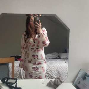 Gina Tricot klänning - Supersnygg långklänning från Gina i vit med rosa och lila blommor. Djup v-ringning, långa puffärmar och knappar framtill hela vägen ner. Använder aldrig den och därför säljer jag, inga fläckar utan som ny då den bara använd en gång💕storlek 34.