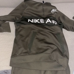 Nike Air hoodie i mörkgrön - Nike Air hoodie i mörkgrön med stor vit logga över bröstet och mindre logga på bröstet. Tröjan har lång ärm, känguruficka och half zip-dragkedja upptill. Perfekt för en sportig och avslappnad stil. Kommer även med byxorna