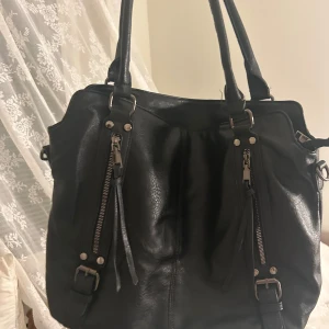 Svart axelväska i skinn med dragkedjor - Snygg svart axelväska i skinn med två långa handtag, dekorativa spännen och dragkedjor framtill. Väskan har även en praktisk ficka med dragkedja på baksidan och coola detaljer i metall. Perfekt för dig som vill ha en stilren och rymlig väska. 