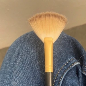 Sminkborste i solfjäderform - En sminkborste med solfjäderformad borst, perfekt för highlighter eller att svepa bort ögonskuggsfall. Borsten har ljust beige strån och ett gult skaft med svart handtag. Enkel att använda för en flawless finish.