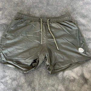 Gröna Moncler badbyxor - Snygga mörkgröna shorts från Moncler med elastisk midja och snörning. Tillverkade i ett lätt syntetmaterial med meshfoder, perfekt för sommardagar. Klassisk Moncler-logga på benet och sportig vibe. Passar dig som gillar stilrena och bekväma plagg. Badbyxor i storlek M. Hör av er vid frågor. Nypris: 3475kr