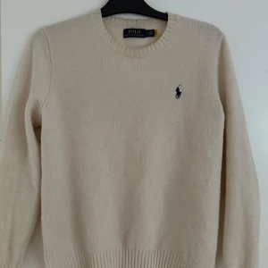 Beige stickad tröja från Polo Ralph Lauren - Säljer en klassisk beige stickad tröja från Polo Ralph Lauren med rund halsringning och den ikoniska broderade loggan på bröstet. Perfekt för lager-på-lager och enkel att matcha med det mesta.
