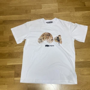 Vit Palm Angels t-shirt med nalletryck - Vit t-shirt från Palm Angels med ikoniskt tryck av en dekapiterad nallebjörn på bröstet och svart logga under. Klassisk passform med rund hals och korta ärmar. Materialet är mjuk bomull som känns skönt mot huden. Perfekt för dig som gillar streetwear med edge.