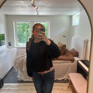 Svart fluffig topp med öppen rygg - Svart fluffig topp med lång ärm och öppen rygg som knyts med ett band i nacken. Materialet har tygdetaljer som ger en unik look. Perfekt för både vardag och fest. Använd 1-2 gånger. 