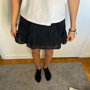 Svart kortkjol från MNG med spets - Supersöt svart kortkjol från MNG med tunna plisserade detaljer och inslag av spets längs sidorna. Kjolen är luftig och har en lätt volang nertill, perfekt för dig som gillar en feminin och chill stil.