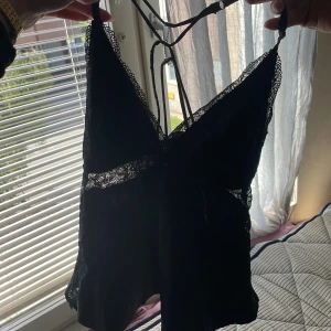 Svart spetsig linnetopp med rosett - Säljer en svart linnetopp med tunna axelband och v-ringning. Toppen har fina spetsdetaljer längs kanterna och under bysten, samt en liten rosett framtill. Materialet är mjukt och glansigt, vilket ger en lyxig känsla. Perfekt för dig som gillar en feminin och trendig stil.