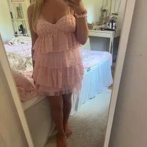 Rosa volangklänning med broderade blommor - Supersöt rosa kortklänning med flera lager volanger och tunna axelband. Klänningen har broderade blommor på tyget och en lätt, luftig känsla. Perfekt för sommarens alla tillfällen. Dragkedja i sidan och justerbara axelband för bästa passform.