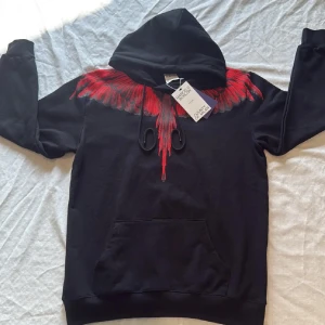 Marcelo Burlon svart hoodie med röd print - Svart hoodie från Marcelo Burlon County of Milan med ikoniskt rött vingmönster över axlar och rygg. Tröjan har huva med snörning och stor magficka framtill. Perfekt statement-plagg för dig som vill sticka ut.
