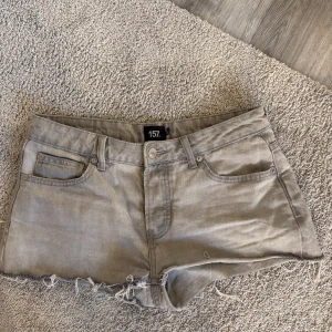 Grå jeansshorts från 157 - Snygga grå jeansshorts från lager 157 med råa, fransiga kanter och klassisk femficksdesign. Shortsen har bälteshällor och knappstängning framtill. Perfekta för en avslappnad och cool stil under varma dagar.