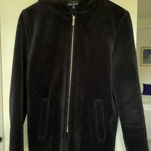 Svart sammetshoodie från Marc O'Polo - Svart hoodiejacka i mjuk sammet från Marc O'Polo. Jackan har dragkedja framtill, huva och två diskreta fickor. Enkel och clean design som passar kvinnor och män. Långärmad och normal passform.