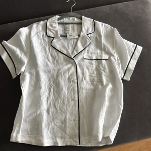 Vit kortärmadpjamas skjorta från H&M - Snygg vit pyjamaskjorta från H&M med svarta kontrastkanter och klassisk krage. Skjortan har korta ärmar, bröstficka och knäppning framtill. Tillverkad i en luftig mix av linne och viskos, perfekt för varma dagar. Aldrig använd pris går att diskutera