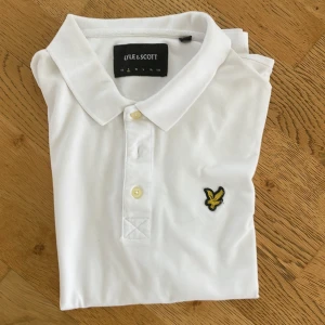 Vit pikétröja från Lyle & Scott S - Klassisk vit pikétröja från Lyle & Scott i storlek S. Tröjan har krage, knäppning med två knappar och den ikoniska gula örnloggan broderad på bröstet. Tillverkad i bomull för en skön könsla☺️ .