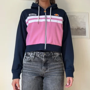 Ellesse croppad zip-hoodie i rosa & marinblå - Snygg croppad hoodie från Ellesse i marinblått och rosa med vita ränder över bröstet. Jackan har dragkedja, huva med snören och broderade Ellesse-loggor framtill. Perfekt för en sportig och chill look.