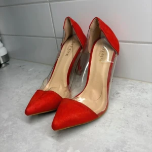 Röda pumps med transparent detalj - Snygga röda pumps från DAY VINE med spetsig tå och höga klackar. Skorna har en cool transparent plastdetalj på sidorna och mockaliknande material på tå och häl. Perfekta för dig som vill sticka ut med en trendig och modern look. Behöver lite uppfräschning.