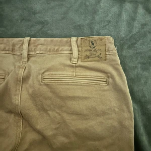 Vintage Beiga jeans från Ralph Lauren - Snygga beiga vintage byxor från Ralph Lauren med förstärkta partier på insidan av benen och dragkedjor på fickorna framtill. Klassisk design med bälteshällor och raka ben. Perfekta för dig som gillar stilrena och funktionella byxor.