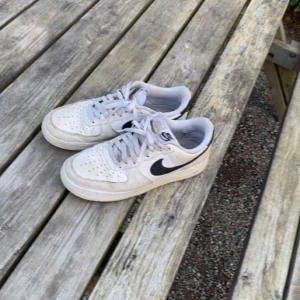 Nike Air Force 1 vita sneakers - Klassiska Nike Air Force 1 sneakers i vitt med svart swoosh. Skorna har snörning, perforerad tå och platt sula. Tillverkade i skinn och syntet, perfekt för dig som gillar streetstyle och tidlös design.