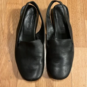 Svarta skinnsandaler från Hobbs - Stilrena svarta sandaler från Hobbs i äkta skinn med fyrkantig tå och öppen häl. Snygg minimalistisk design med bred rem över foten och klassisk slingback. Perfekta för dig som gillar en clean och tidlös look.