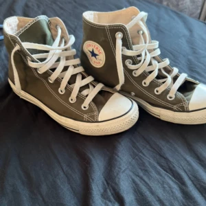 Olivgröna Converse All Star high - Snygga olivgröna Converse All Star high sneakers med klassisk vit sula och tå, vita snören och tydliga loggor på sidan och hälen. Skorna är i canvas och har rund tå och platt sula. Perfekta för dig som gillar en clean och tidlös streetstil.