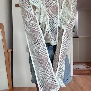 White lace top - Superfin vit blus i spets med lång ärm och volangdetaljer på axlarna. Blusen är transparent med ett unikt geometriskt mönster och hög hals. Perfekt för dig som vill sticka ut med en romantisk och trendig look.