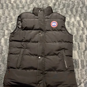 Svart dunväst från Canada Goose - Säljer en svart dunväst från Canada Goose med klassisk logga på bröstet och broderad etikett på ryggen. Västen har hög krage, tryckknappar framtill och två fickor med dragkedja. Perfekt för lager-på-lager och riktigt snygg till streetwear.