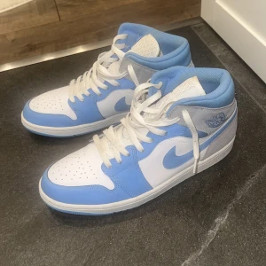 Nike Air Jordan 1 Mid UNC blå/vit - Nike Air Jordan 1 Mid i fräsch UNC-färg med ljusblå och vita paneler samt grå detaljer i mocka. Klassisk Jordan-logga på sidan, perforerad tå och vit snörning. Skön vadderad krage och ikonisk swoosh på sidorna. Perfekta för dig som vill sticka ut med stil.
