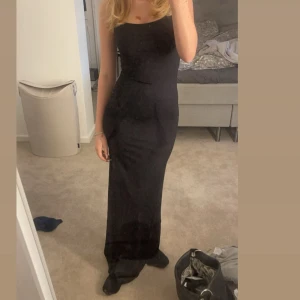 Svart långklänning med smala axelband - En stilren svart långklänning med smala axelband och figurnära passform. Klänningen är ärmlös och har en enkel, elegant design som ger en snygg siluett. Perfekt för dig som gillar minimalistisk stil.