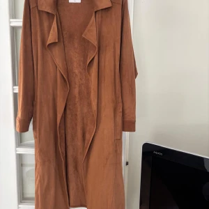 Ljusbrun trenchcoat från Gémo - Snygg ljusbrun trenchcoat från Gémo i mockaimitation. Jackan har långa ärmar, klassisk krage och ett öppet, avslappnat fall framtill. Perfekt för dig som vill ha en stilren och trendig look till vardags.